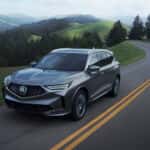 Side view 2026 Acura MDX in grey driving on rural road amongst evergreen trees