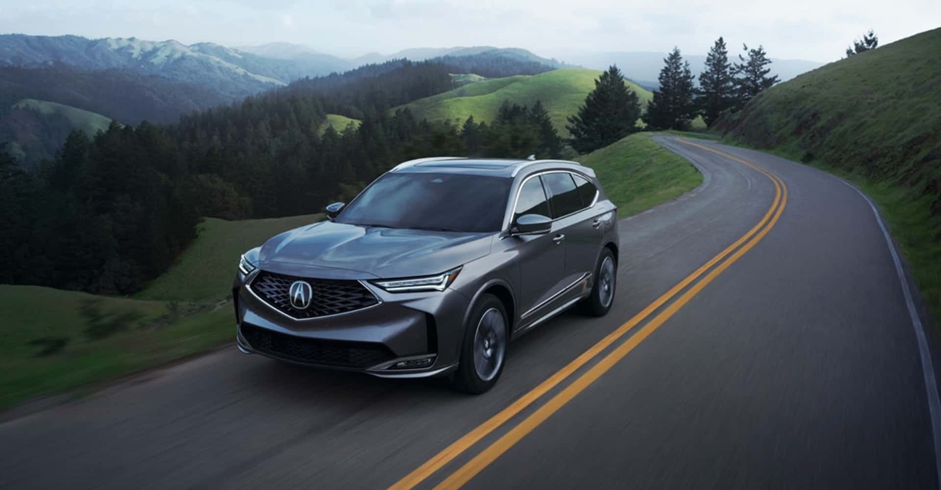 Side view 2026 Acura MDX in grey driving on rural road amongst evergreen trees