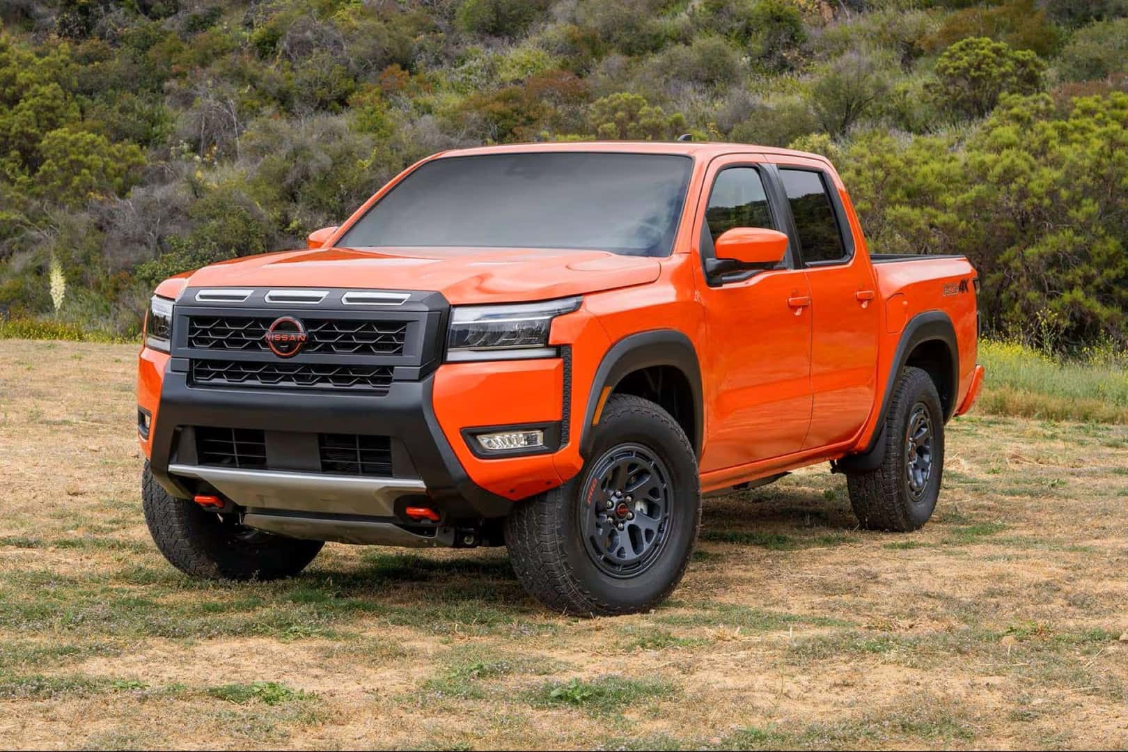 The Competent Nissan Frontier, Parked Effectively In The Middle Of A Grass Field, With A Hill Of Bushes Behind It
