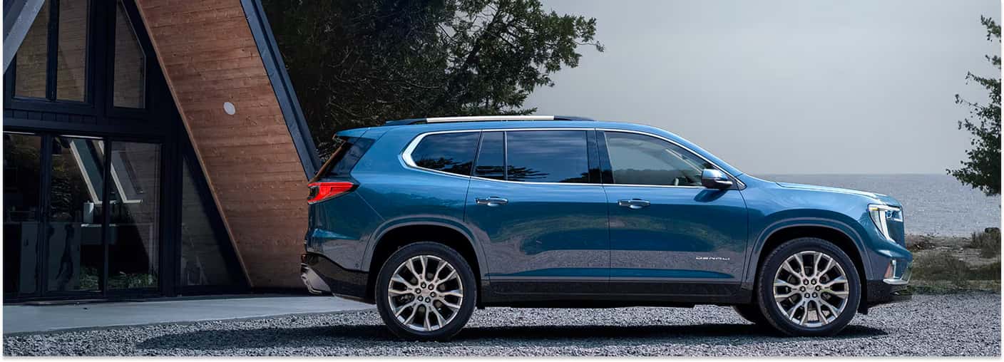 Side profile view of a metallic blue 2026 GMC Acadia Denali parked outside a modern A-frame cabin.