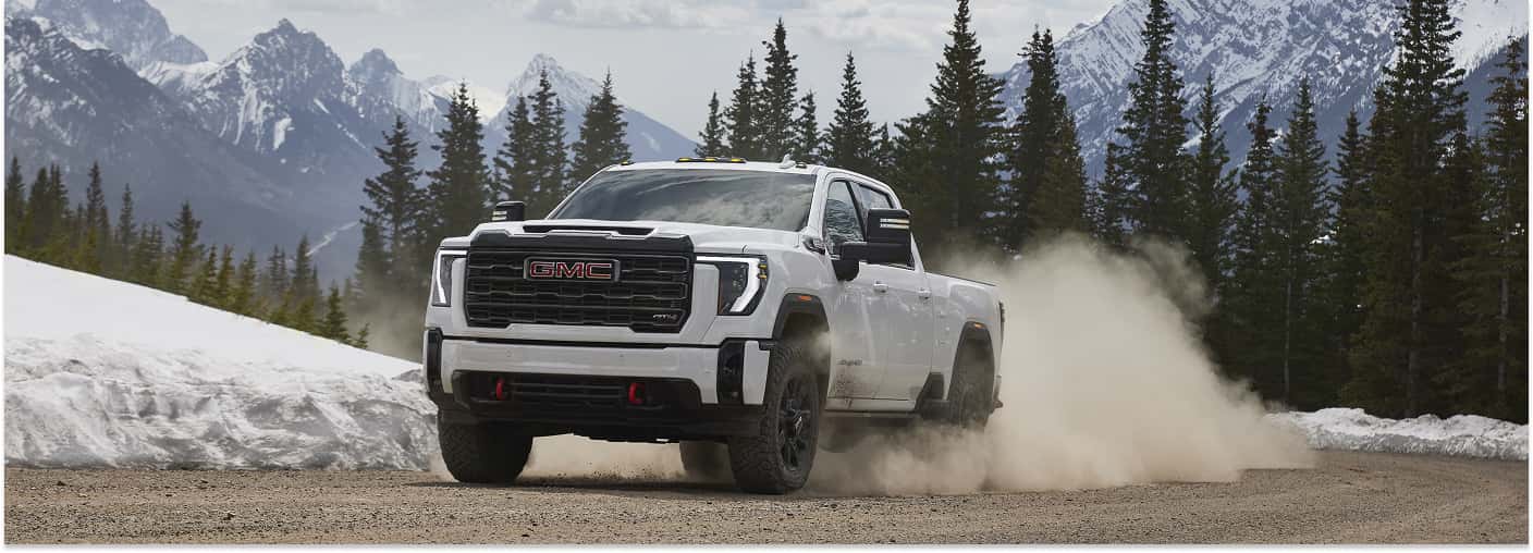 Front view of a white 2026 GMC Sierra 2500 HD AT4 kicking up dirt on a gravel road near snowy mountains.