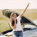 Positive woman using smartphone near broken automobile in countryside during car travel