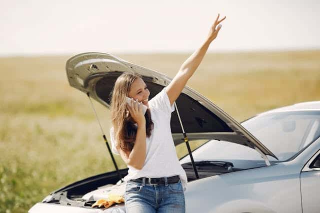 Positive woman using smartphone near broken automobile in countryside during car travel