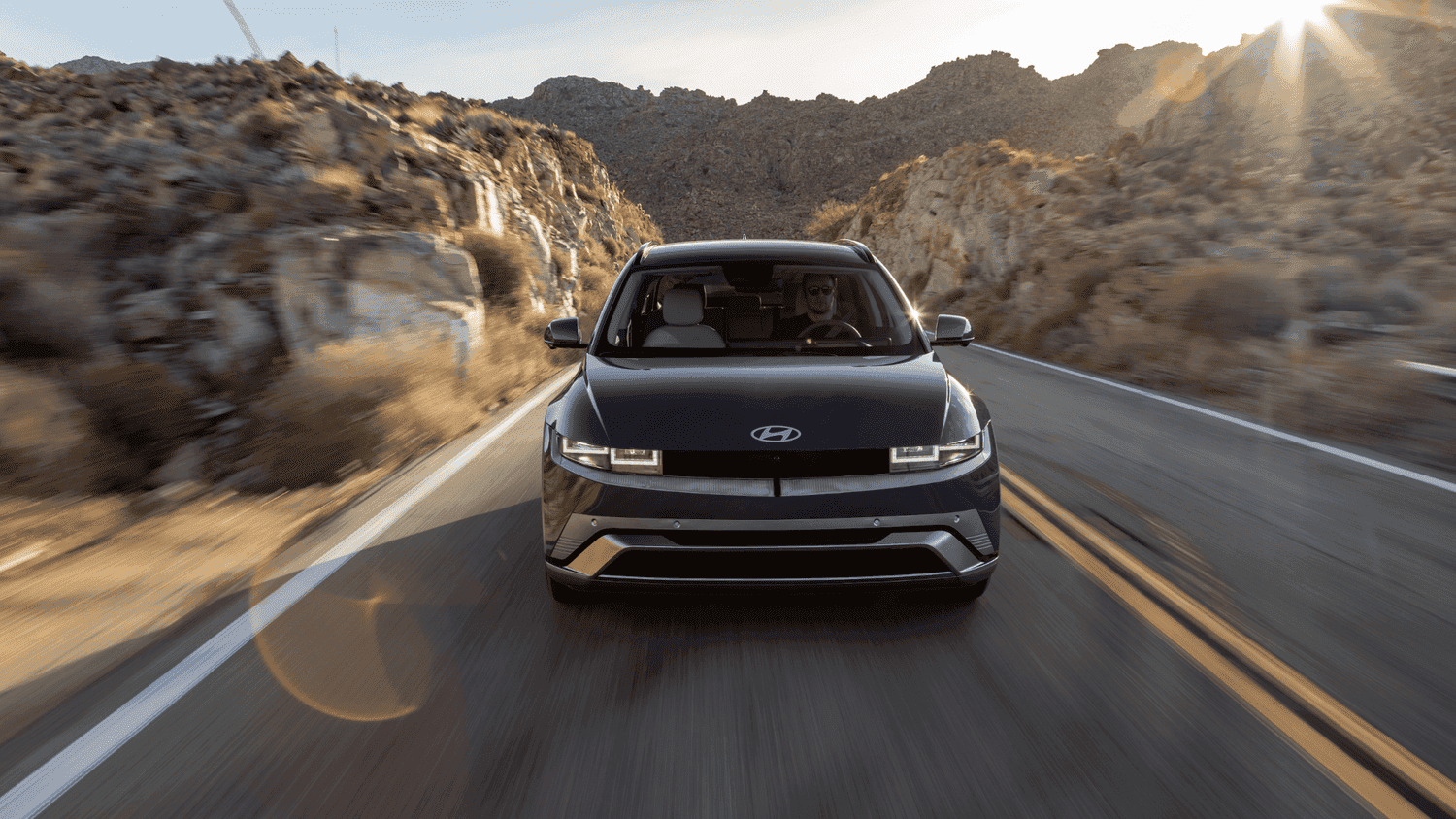 Front view of the 2025 Hyundai IONIQ 5 driving through scenic roads in Athens, GA, with the sun setting in the distance and rugged terrain ahead.