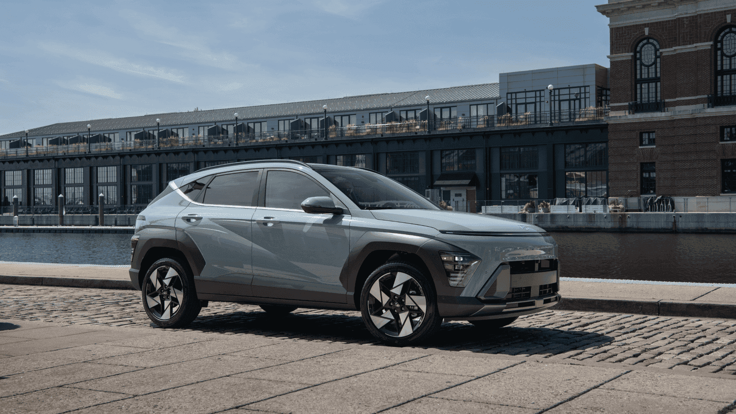Side view of the 2025 Hyundai Kona in Athens, GA, parked near a modern waterfront complex, showcasing its bold urban styling and crossover stance.