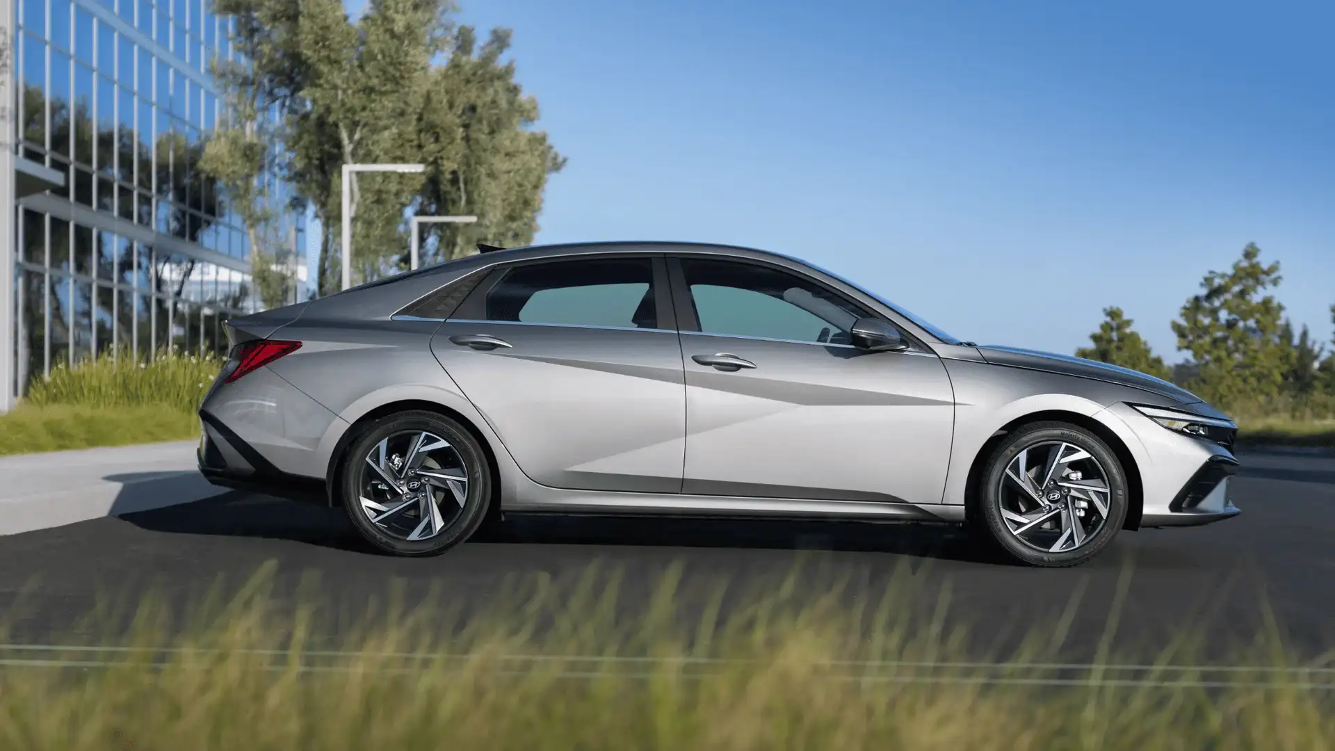 Side profile of silver 2025 Hyundai Elantra parked in front of a glass building with sharp body lines and alloy wheels.