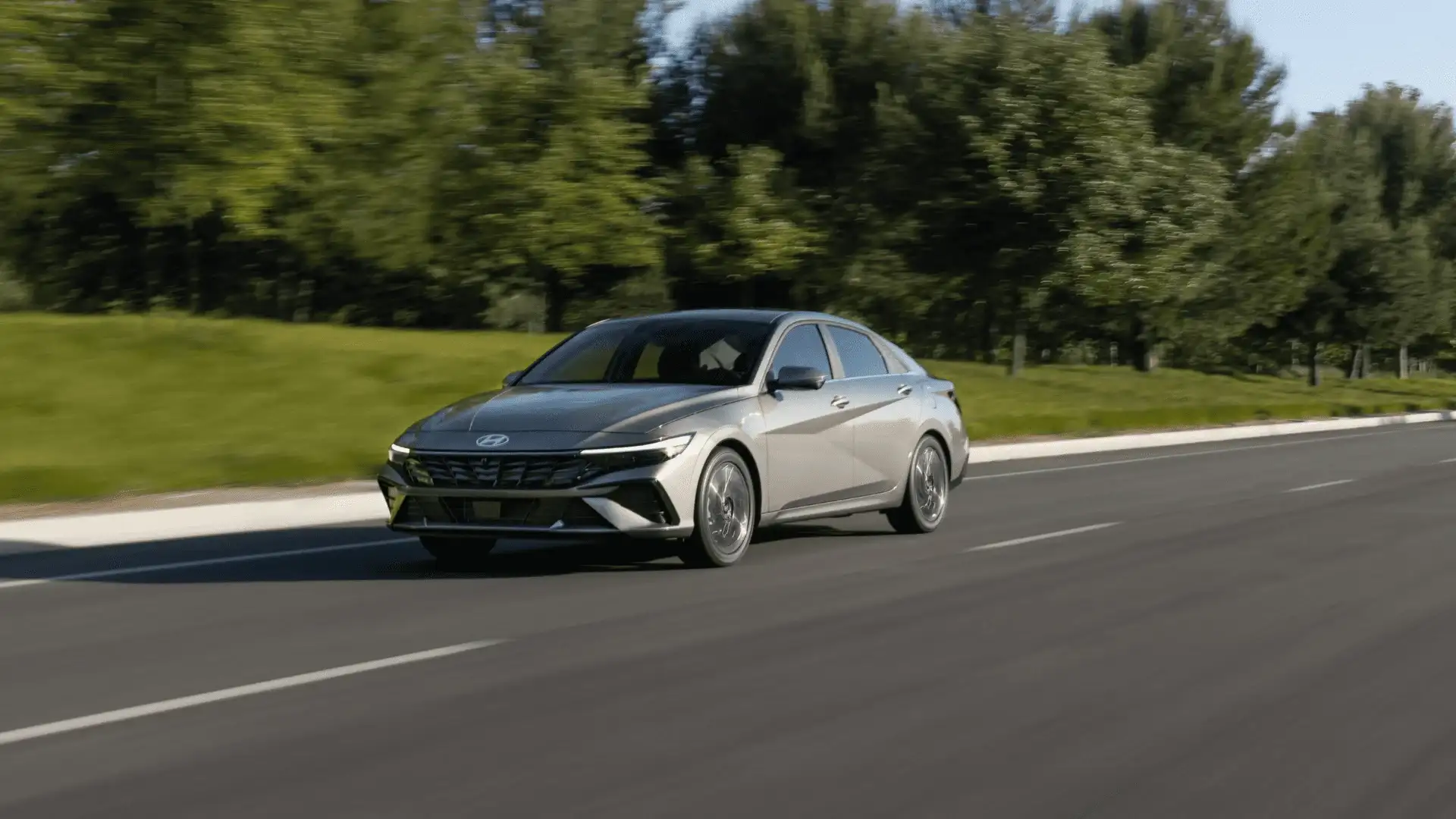 Front three-quarter view of silver 2025 Hyundai Elantra driving on a tree-lined road under daylight.