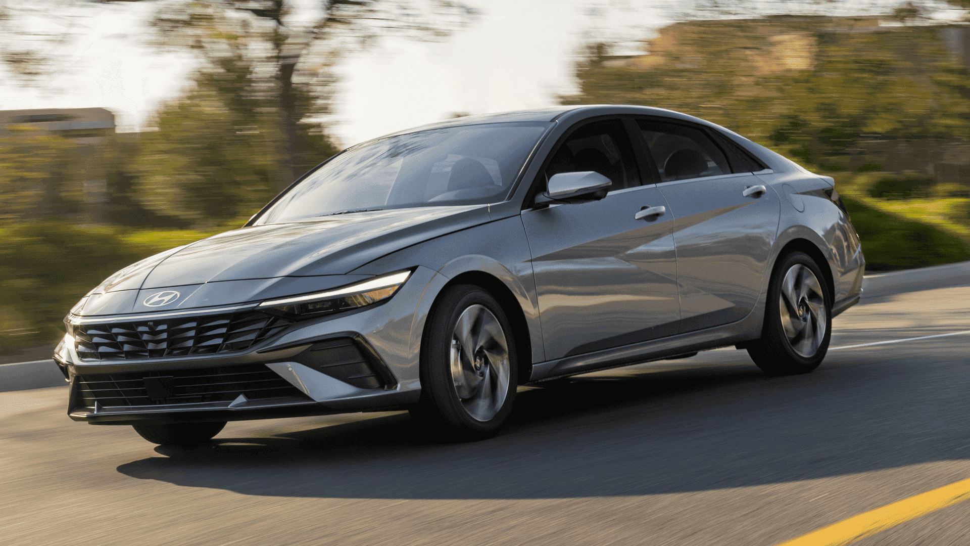 Silver 2025 Hyundai Elantra sedan in motion on suburban road, front-side angle view with LED lights.