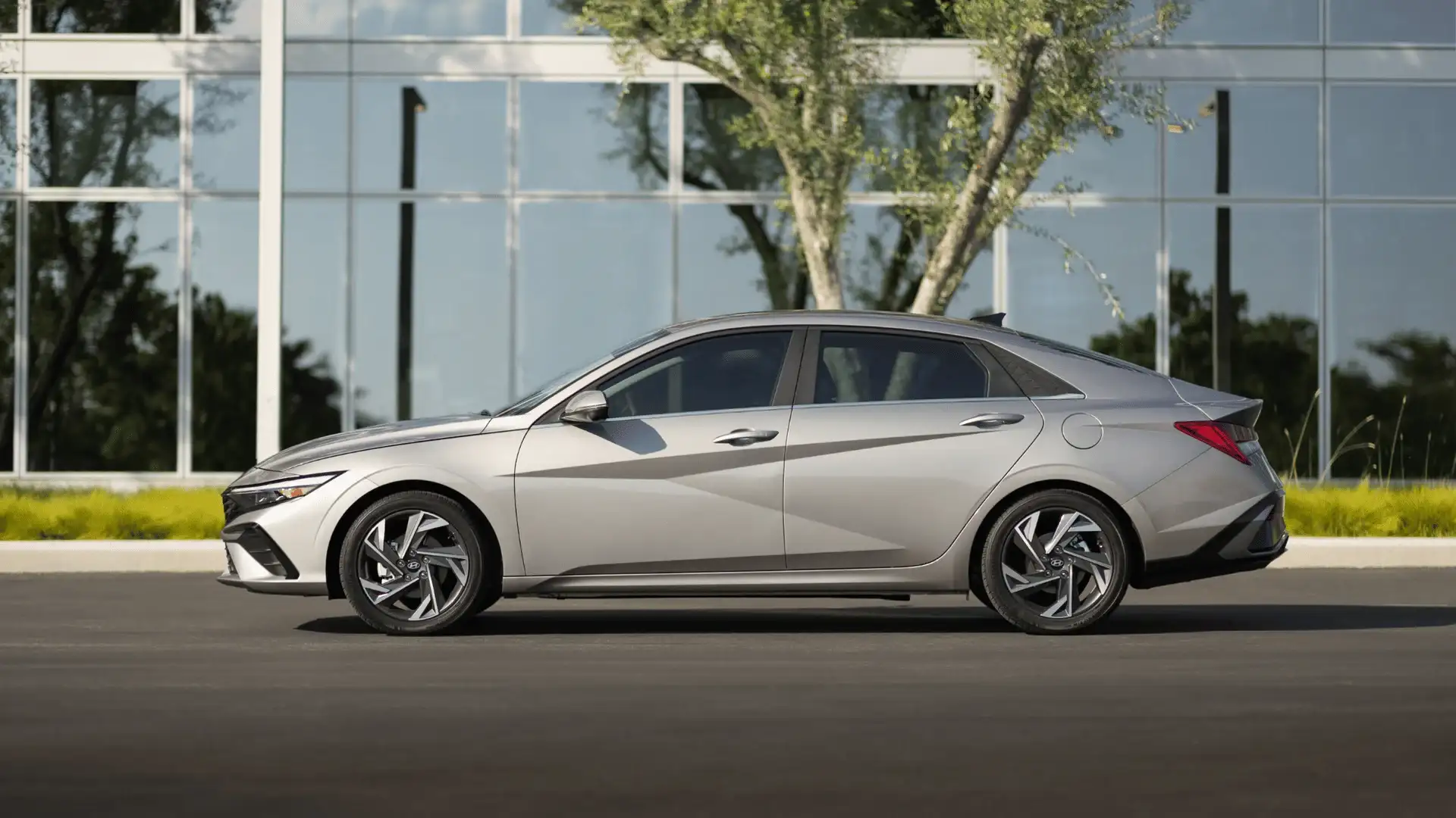 Profile view of a silver 2025 Hyundai Elantra parked in front of a glass office building with modern landscaping..