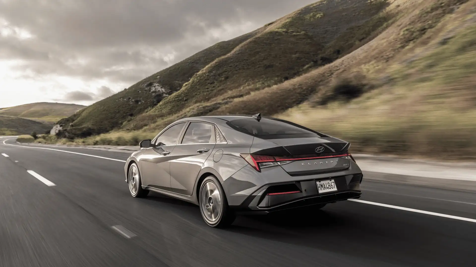 Rear-angle driving shot of a 2025 Hyundai Elantra on a highway, showing its sculpted taillights and sloping fastback design.