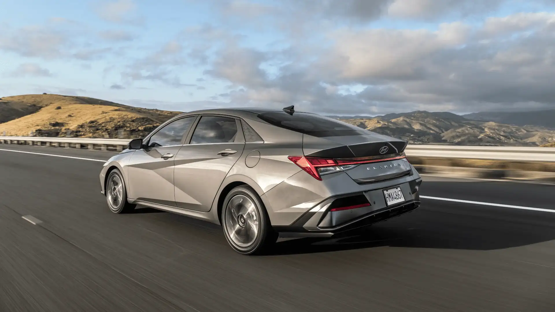 Rear-side view of 2025 Hyundai Elantra driving on a highway, showcasing its aerodynamic tail and LED taillights.