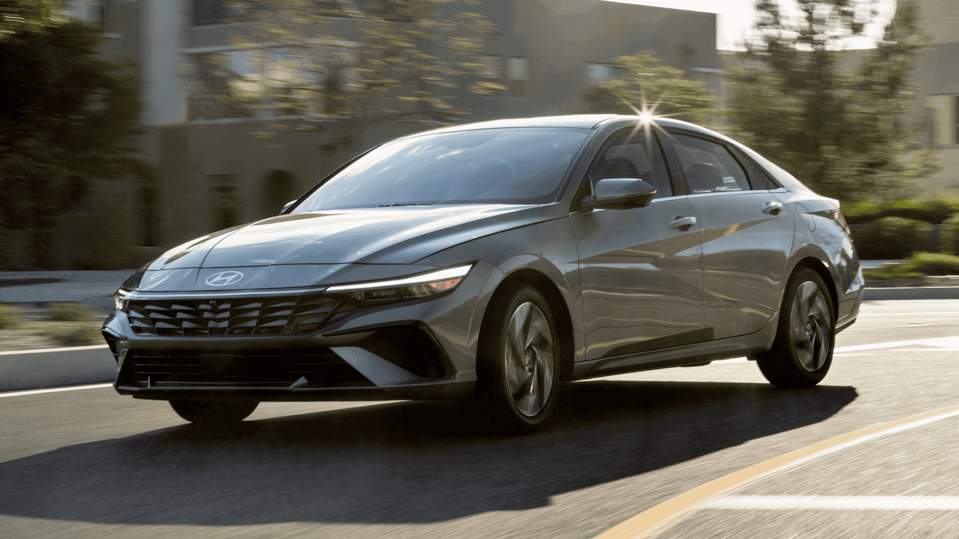 2025 Hyundai Elantra in silver driving on sunlit city street, showing aerodynamic design and wheels.