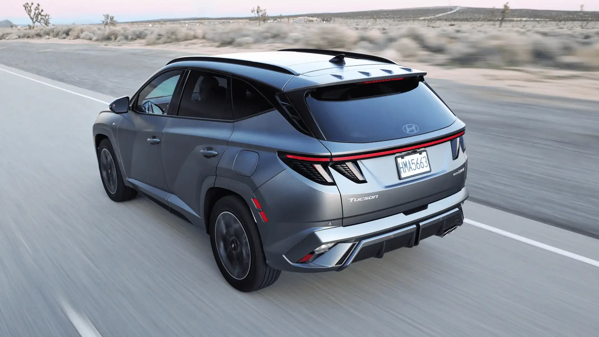 Rear-angle view of the 2025 Hyundai Tucson in gray driving on highway, showing LED taillights and roof spoiler.