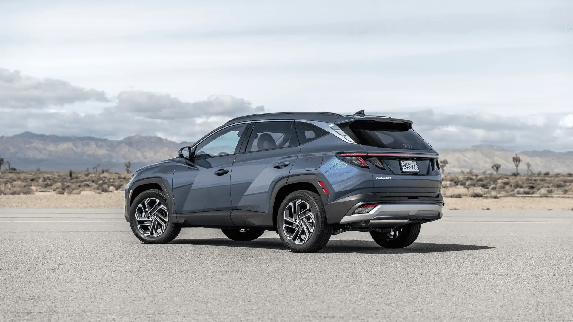 Static rear-side view of 2025 Hyundai Tucson in gray with two-tone wheels, parked against mountain landscape.
