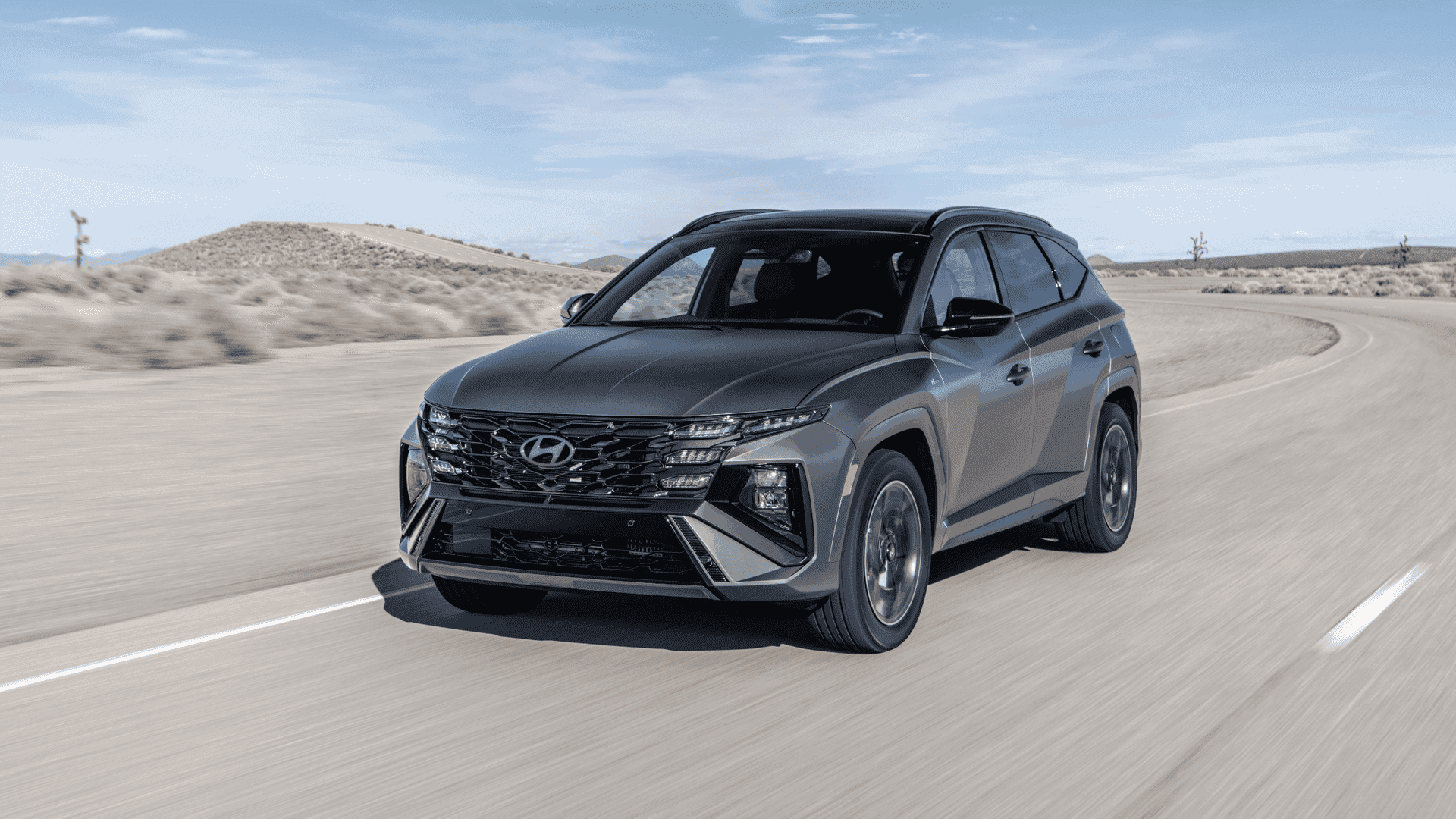Matte black 2025 Hyundai Tucson driving on desert road, close-up front angle with angular grille.