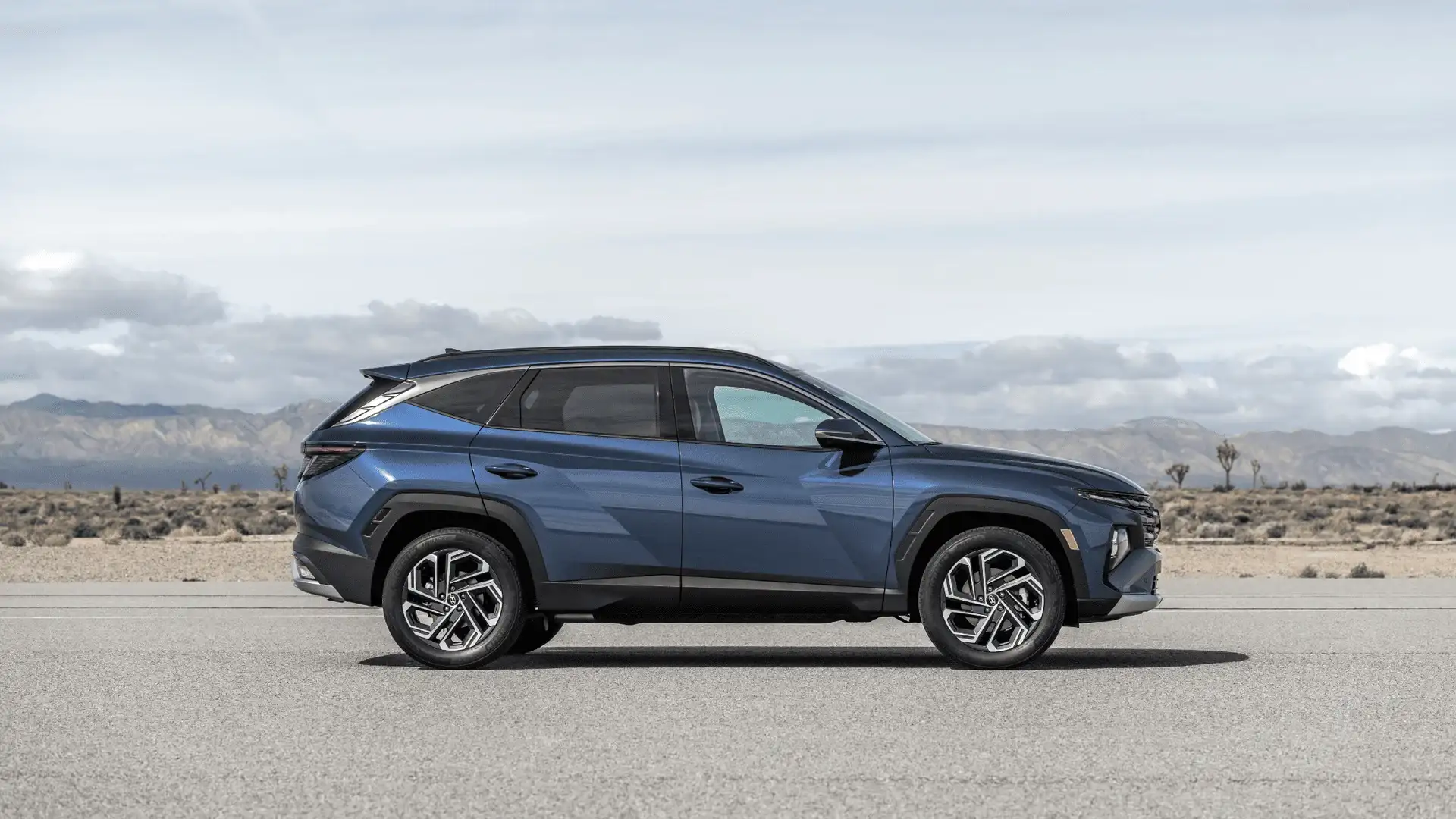Side profile of a blue 2025 Hyundai Tucson SUV parked in a desert landscape with mountains in the distance.