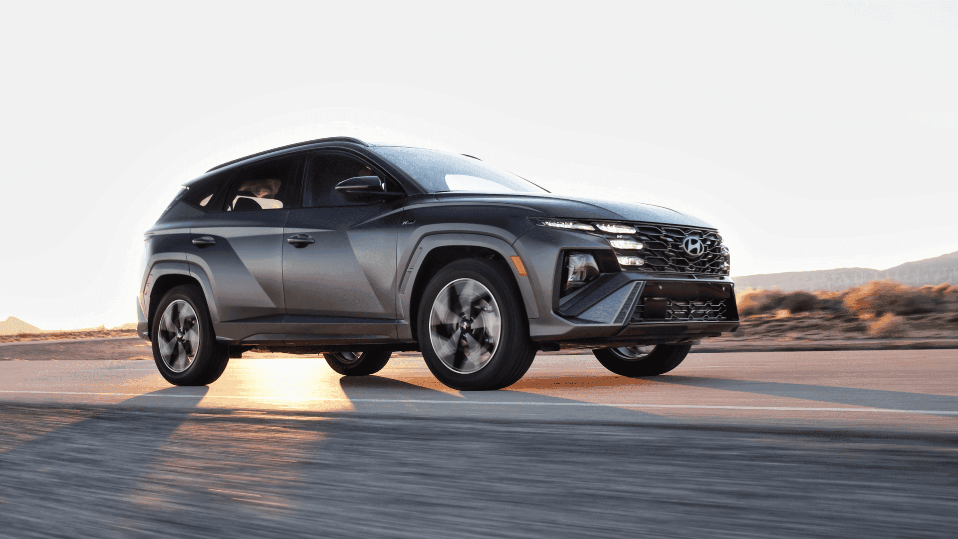 Side profile of 2025 Hyundai Tucson in motion during sunrise, dark exterior with sporty front fascia.