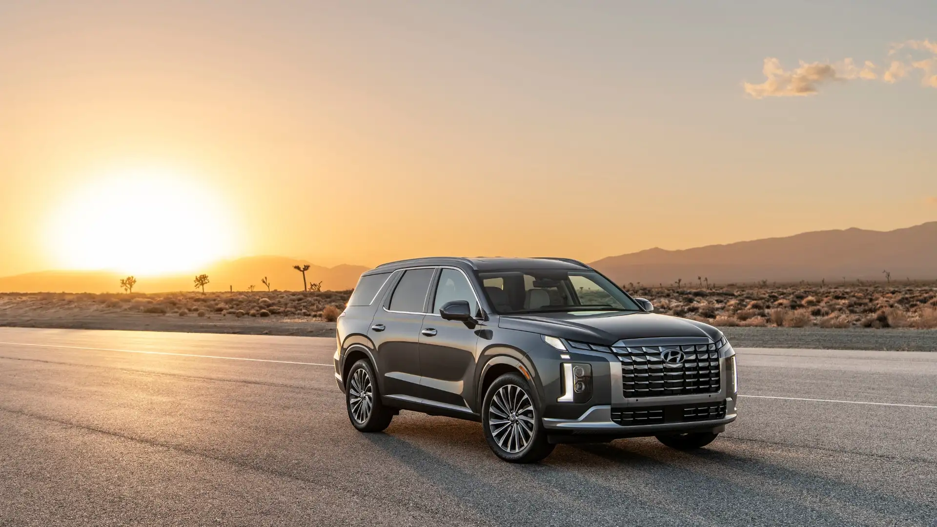 Front three-quarter view of a gray 2025 Hyundai Palisade SUV parked on an open desert road at sunset, showcasing its bold grille, LED lighting, and refined design.