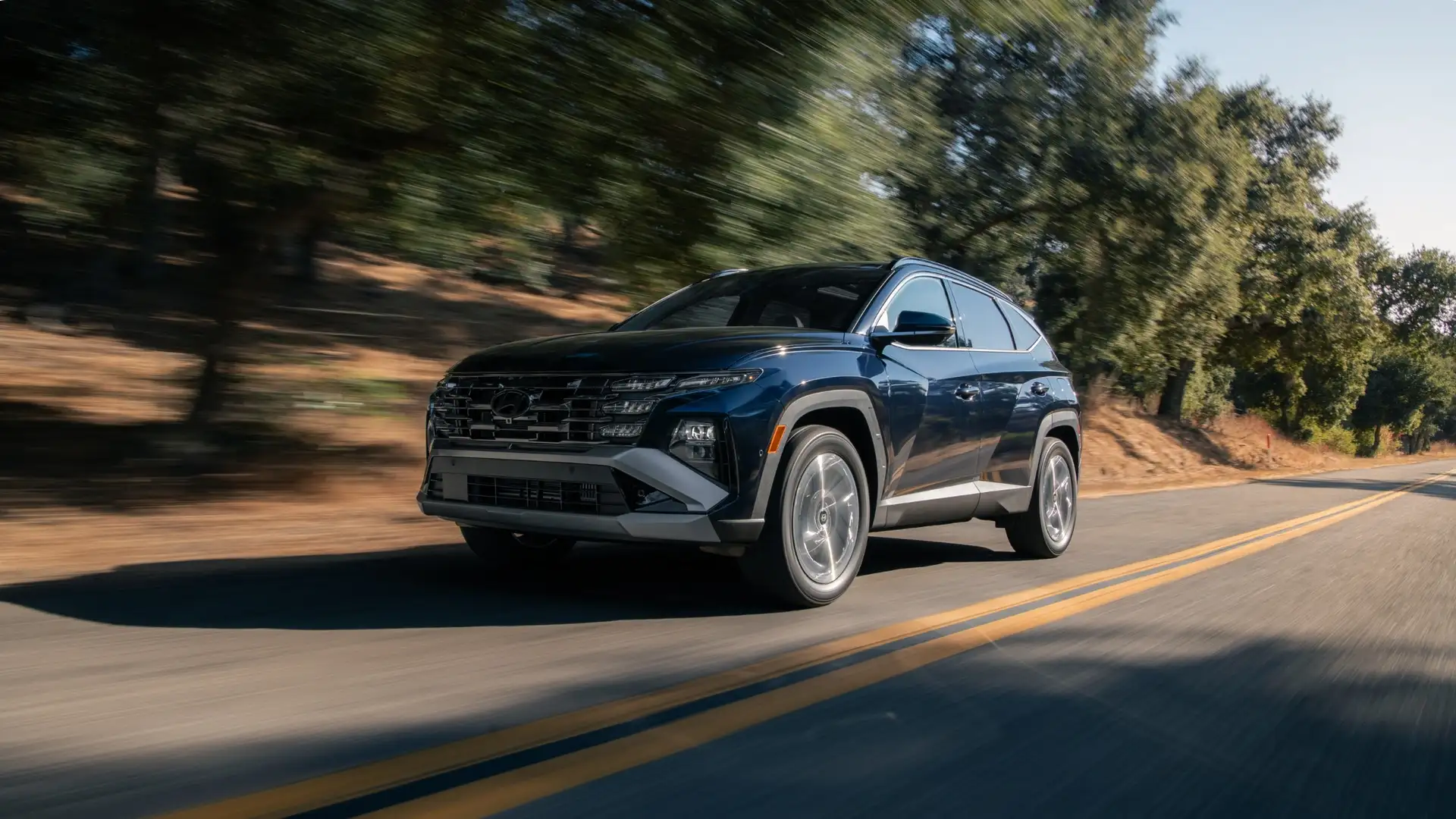 2025 Hyundai Tucson Hybrid SUV driving on a scenic forest road emphasizing fuel efficiency and modern design.