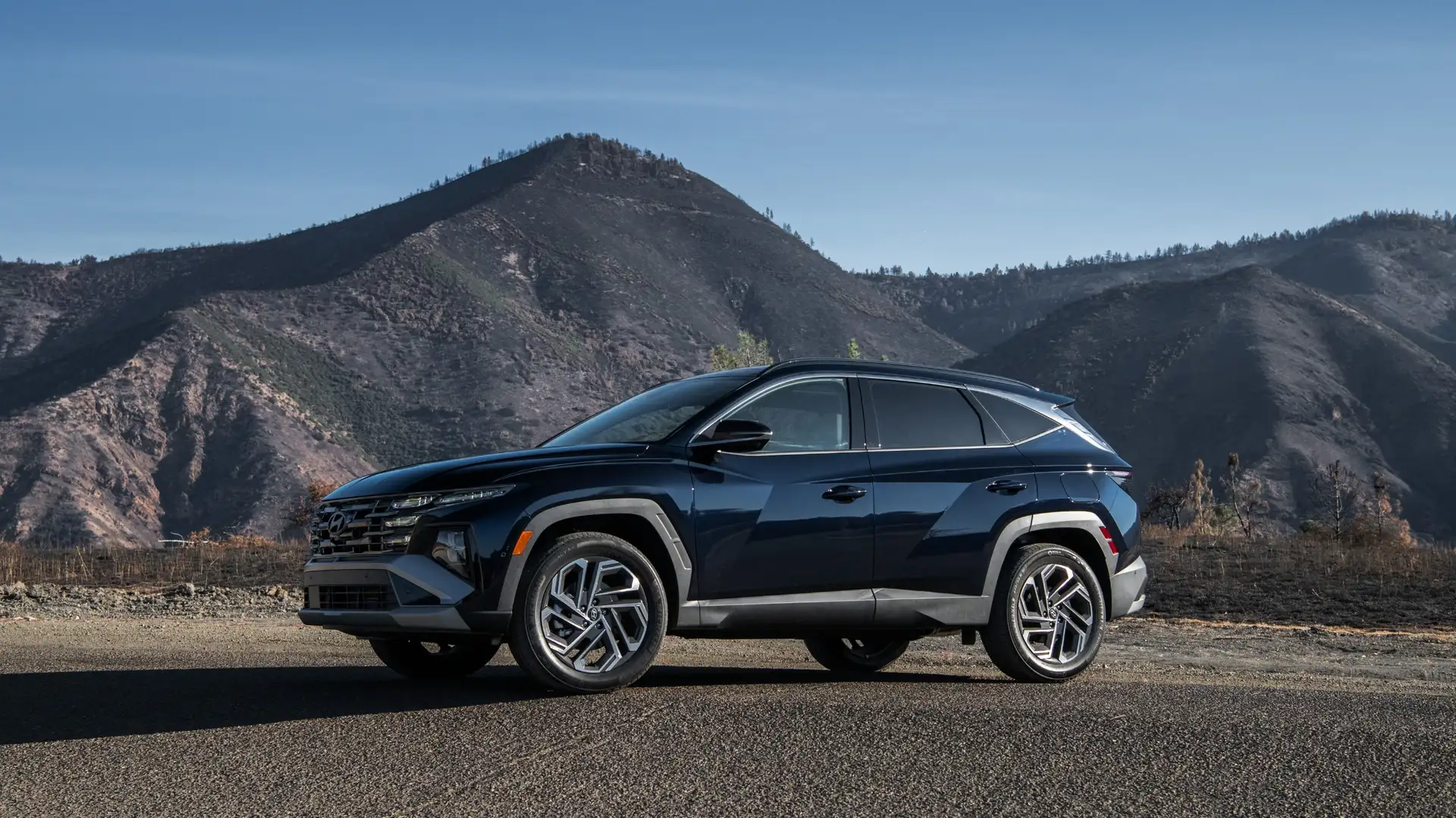 Side profile of a dark blue 2025 Hyundai Tucson Hybrid parked along a scenic mountain road, highlighting its bold geometric body lines and modern SUV design.