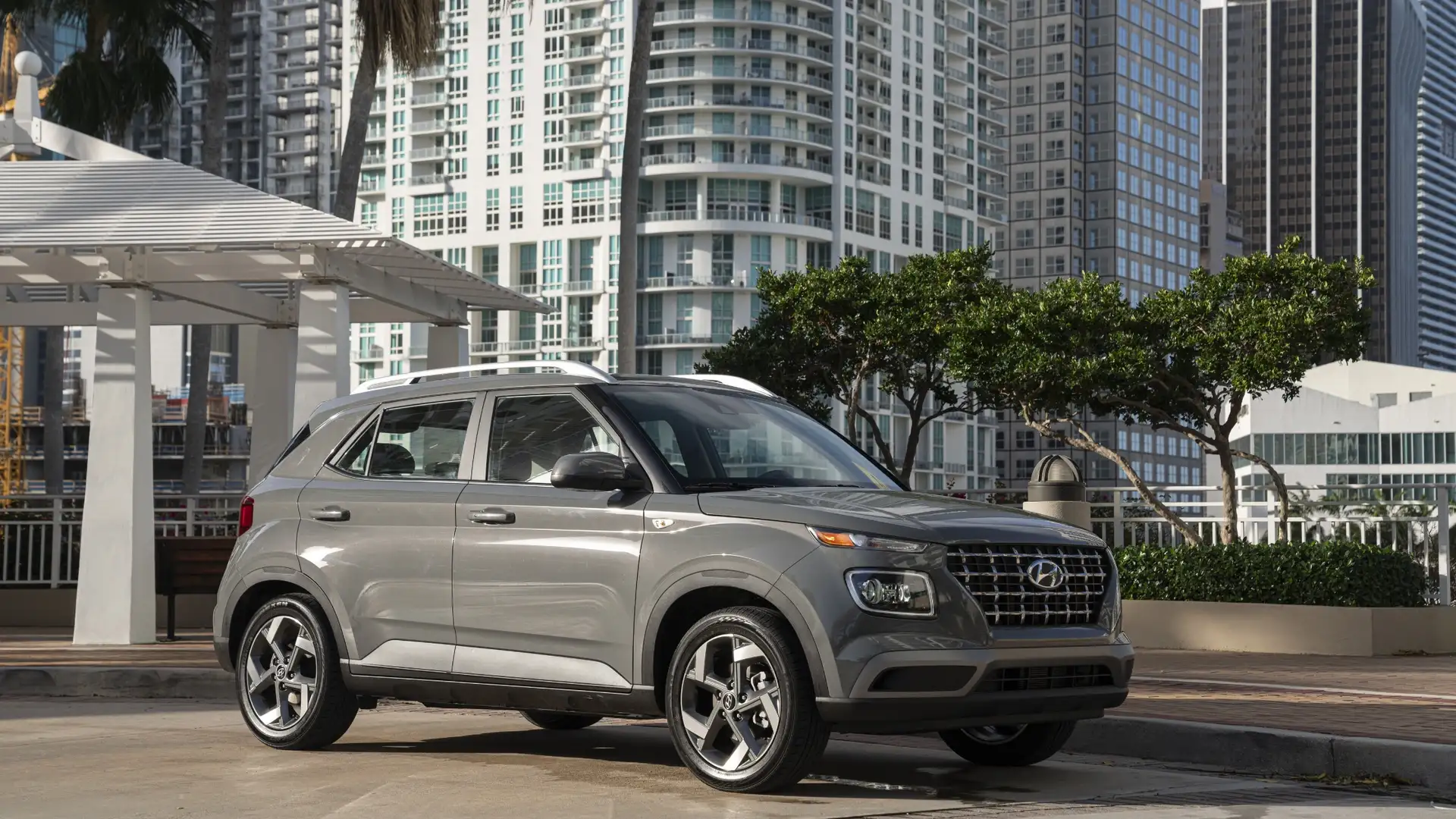 Side profile of a gray 2026 Hyundai Venue compact SUV parked in an urban setting, showcasing its bold front grille, alloy wheels, and modern city styling.