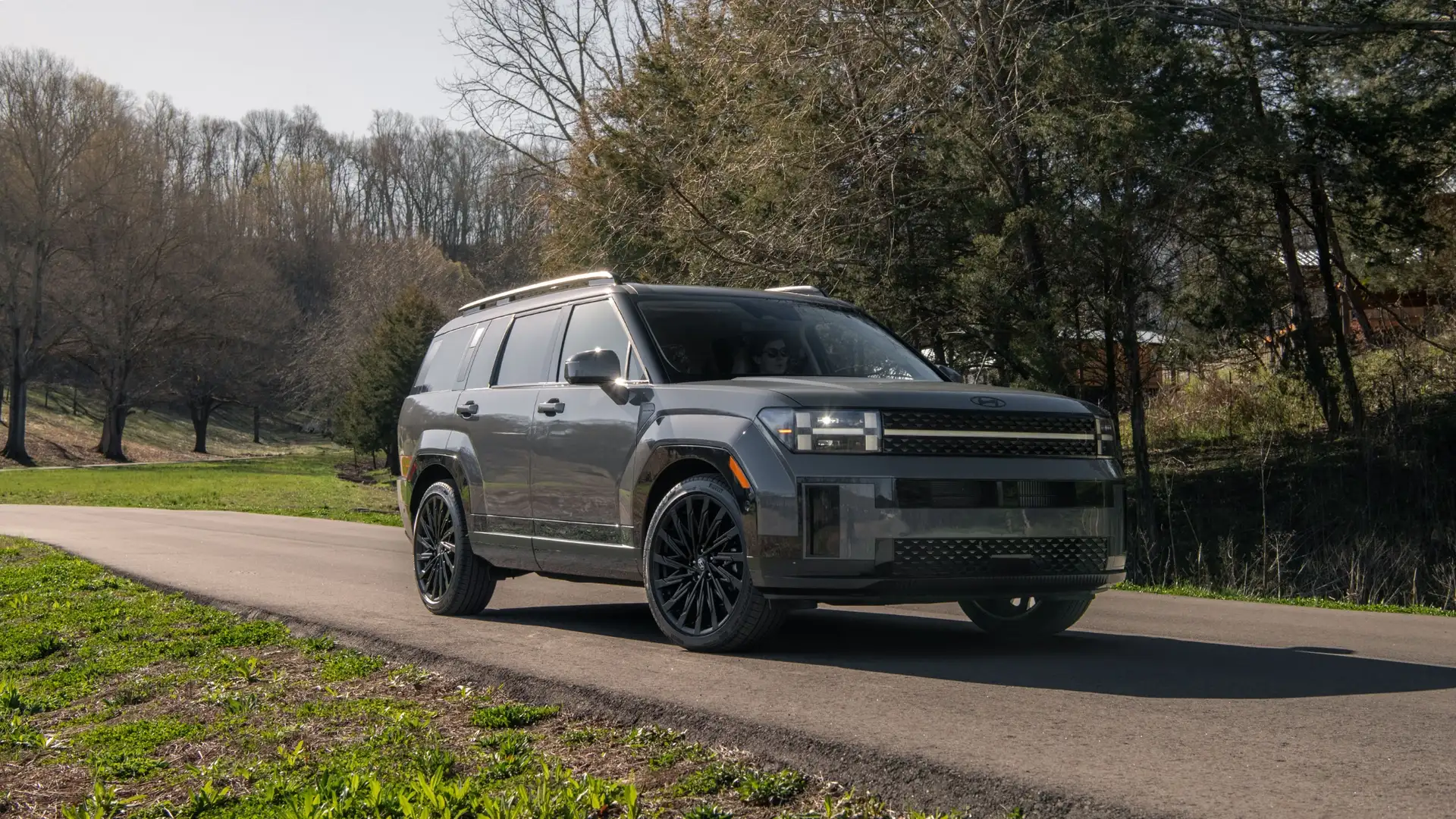 Front-angle view of a 2026 Hyundai Santa Fe in matte gray driving on a rural road, highlighting its bold boxy design and black alloy wheels.