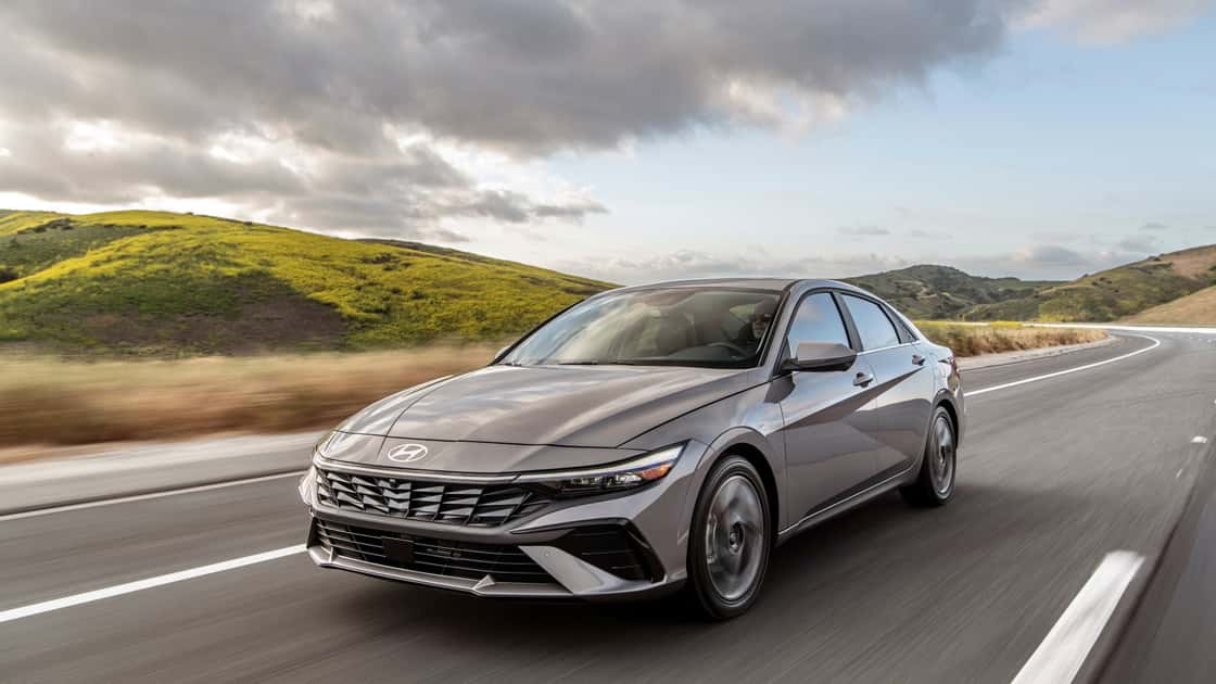 Side-front view of gray 2026 Hyundai Elantra sedan in motion on an open road with scenic hills in the background.