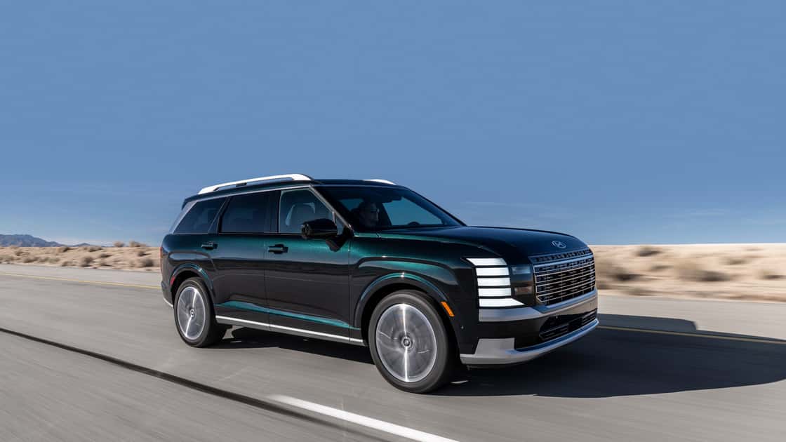 2026 Hyundai Palisade dark green exterior driving on a desert road, showing front grille, headlights, and side profile.