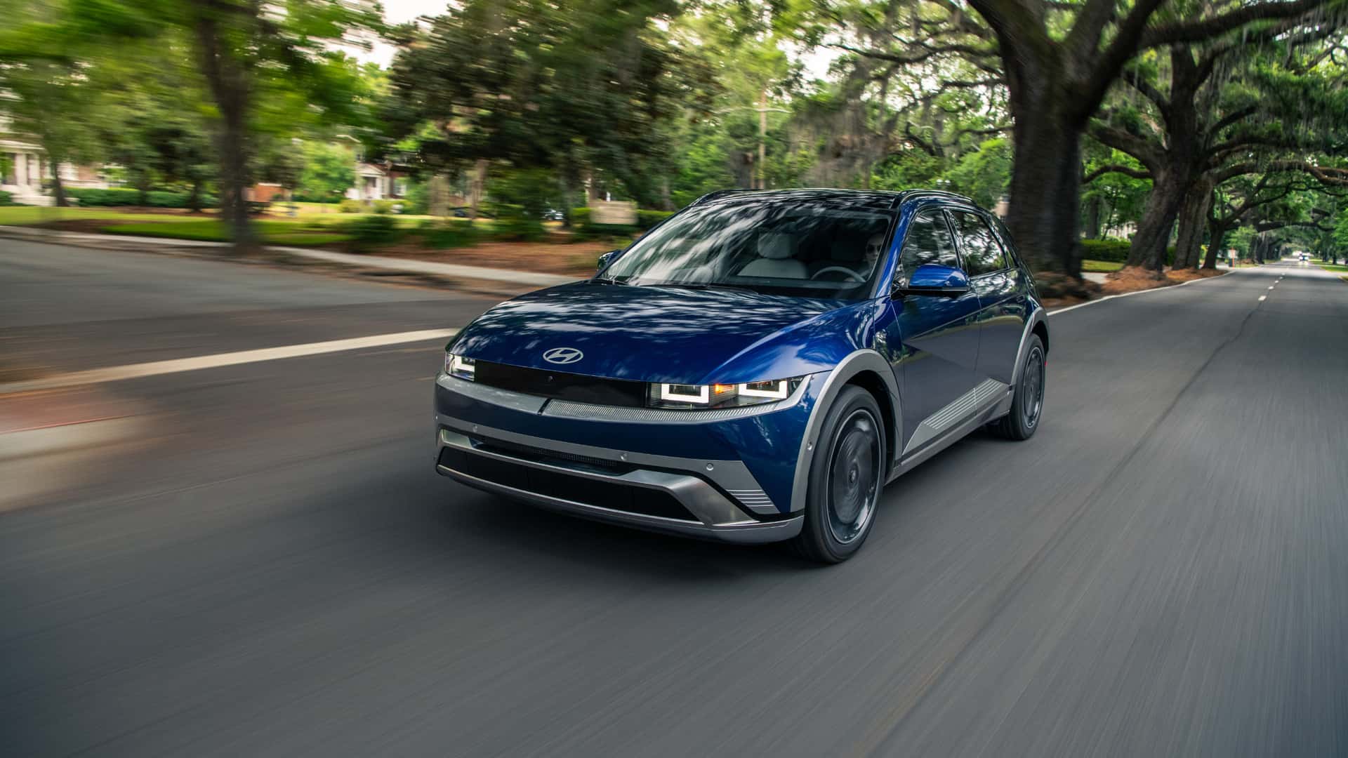 2026 Hyundai IONIQ 5 blue electric SUV driving through a tree-lined street with modern LED lighting.