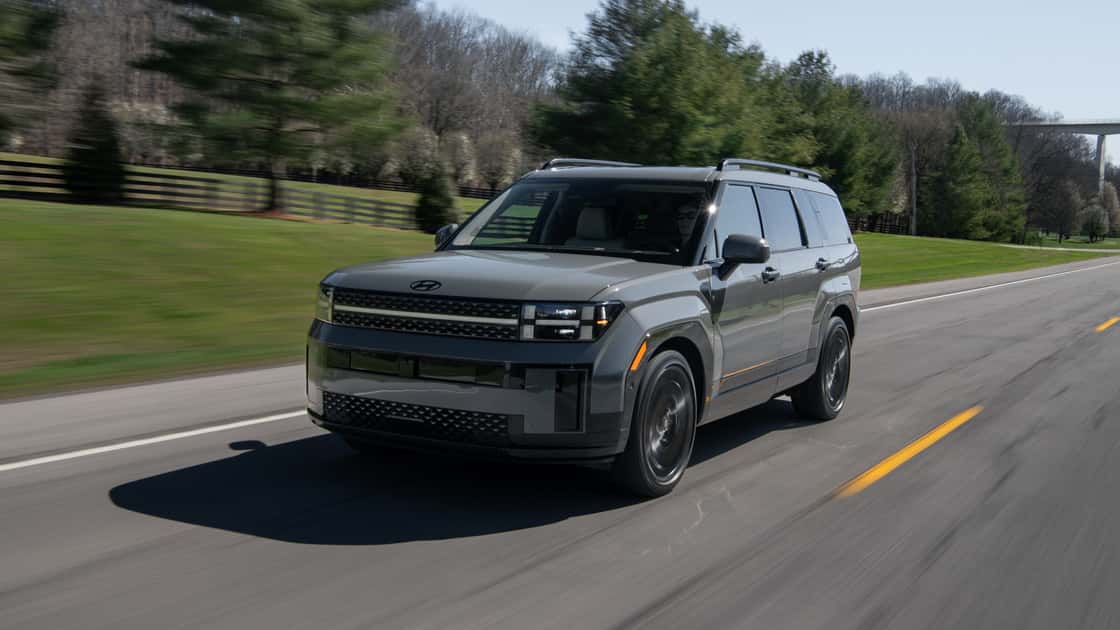 2026 Hyundai Santa Fe gray SUV driving on a rural road with boxy styling, roof rails, and LED lighting.