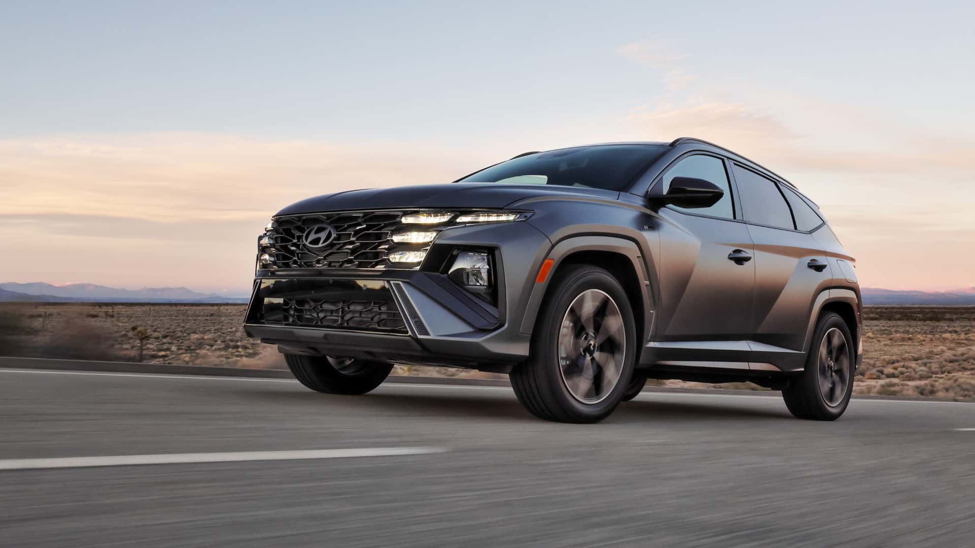 2026 Hyundai Tucson gray SUV driving on a desert highway at sunset with sharp grille design and LED lights.