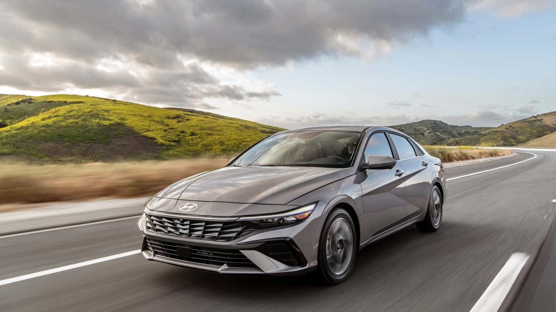 2026 Hyundai Elantra sedan driving on a highway with gray exterior, sharp front styling, and mountain road scenery.