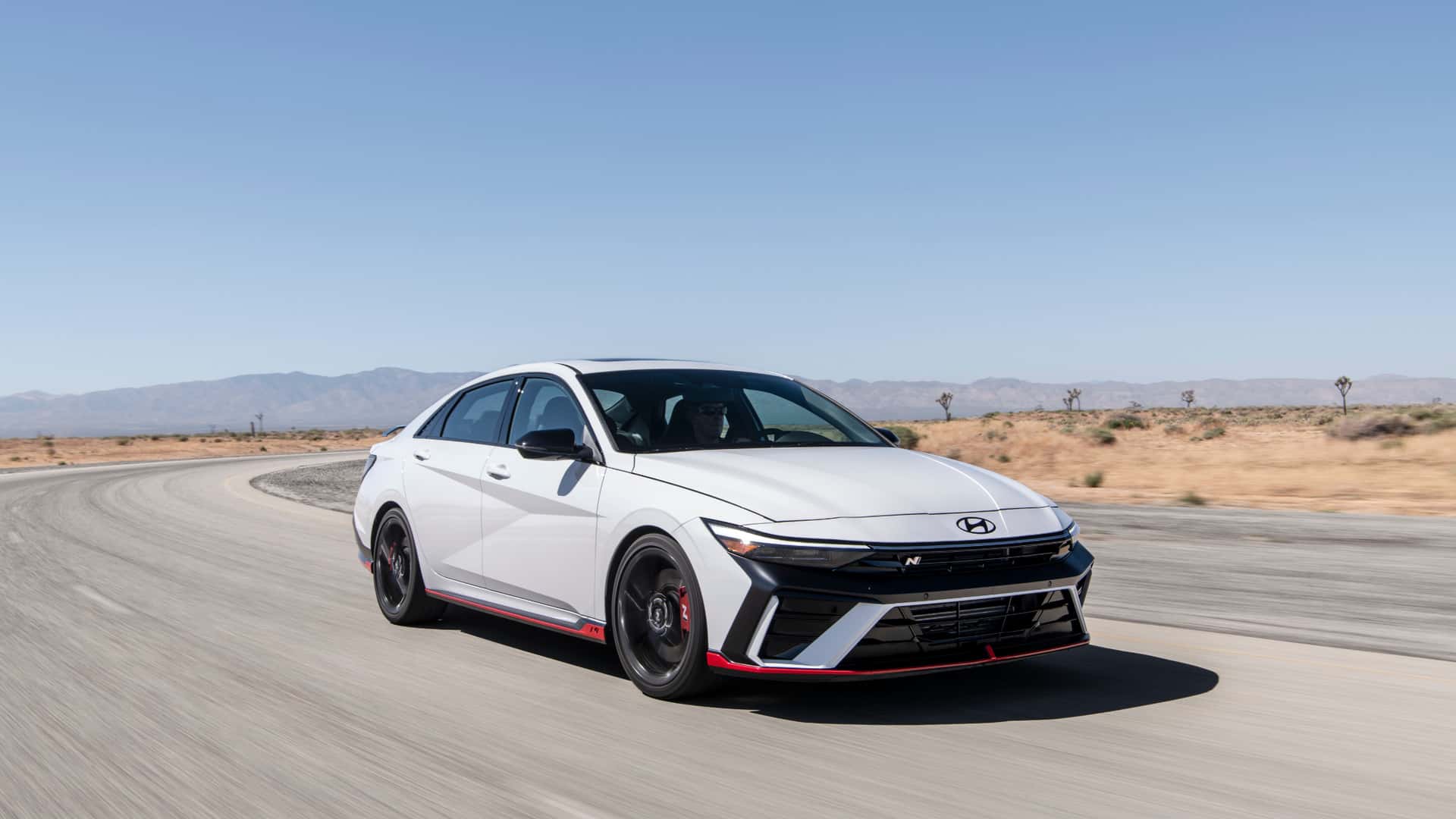 2026 Hyundai Elantra N in white driving on a desert road with black wheels, red accents, and sporty N styling.