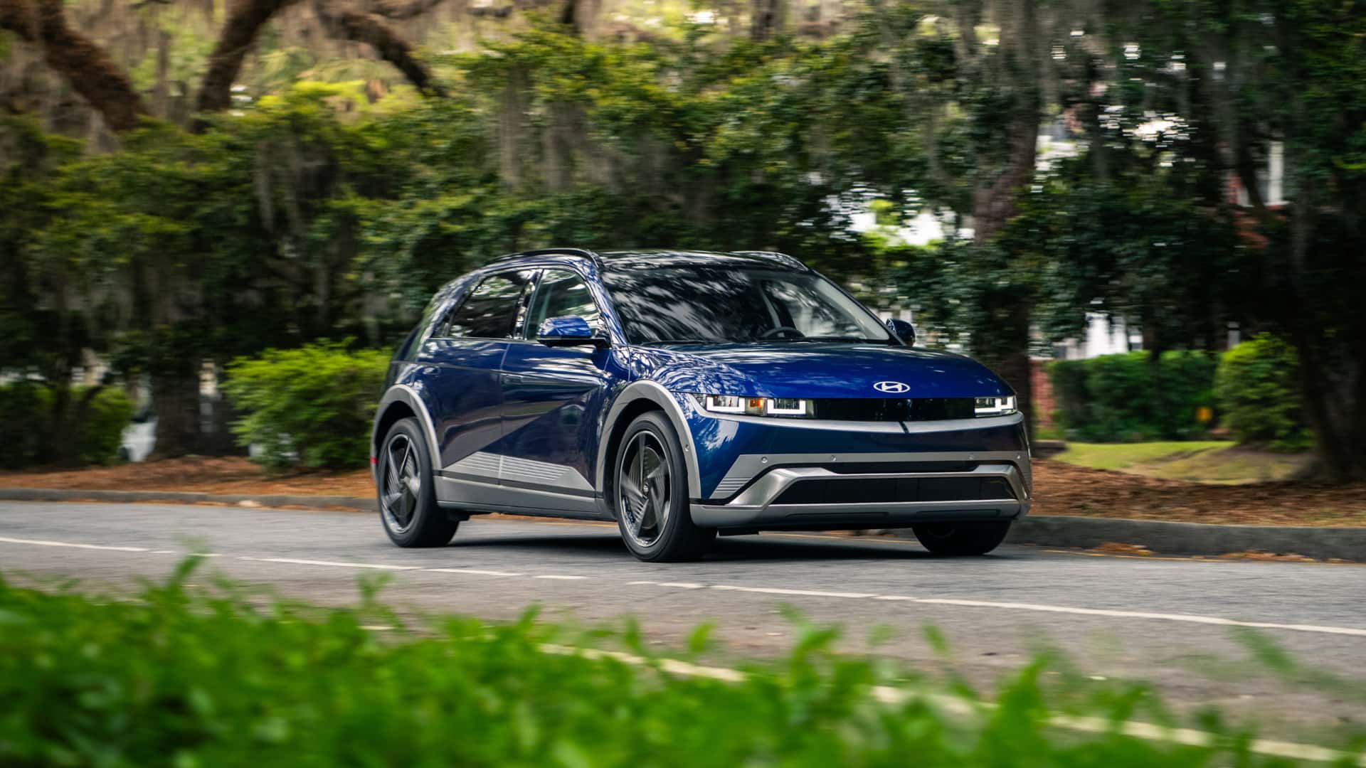 2026 Hyundai IONIQ 5 in blue driving through a tree-lined road with pixel-style headlights and electric SUV design.