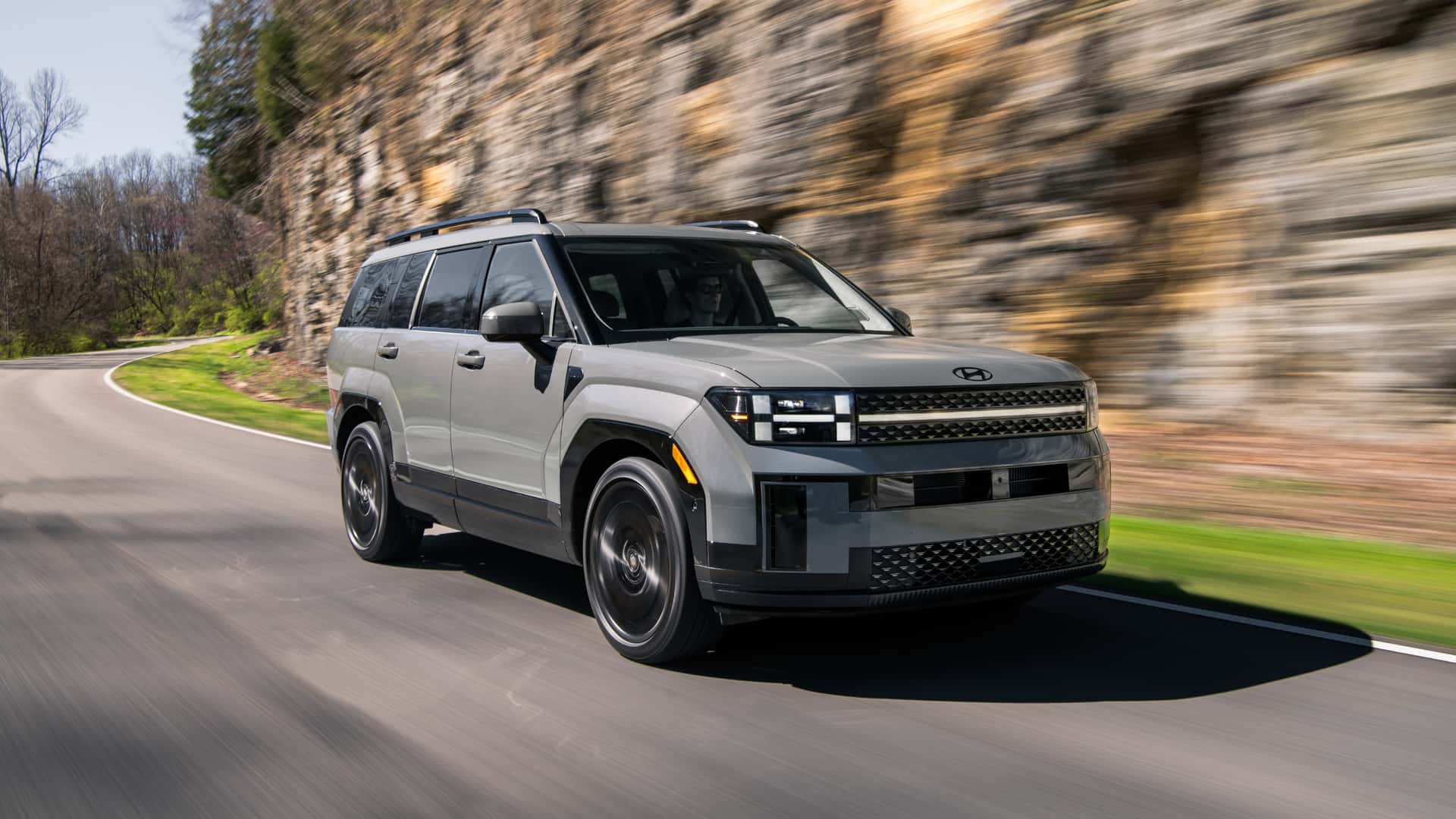 2026 Hyundai Santa Fe gray SUV driving on a winding road with bold grille, roof rails, and rugged boxy styling.