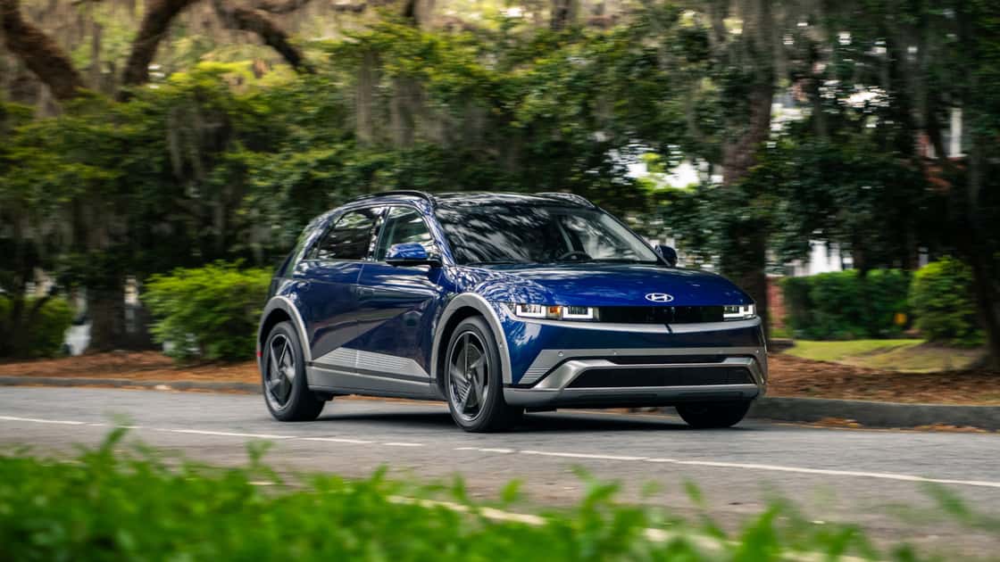 2027 Hyundai IONIQ 5 blue electric SUV driving on a tree-lined road with modern LED lighting and compact crossover styling.