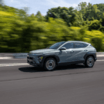 Light green 2025 Hyundai Kona driving on highway with forest blur in the background.