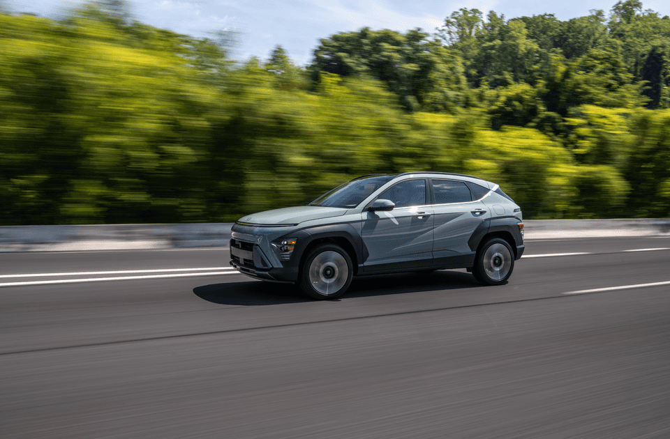 Light green 2025 Hyundai Kona driving on highway with forest blur in the background.