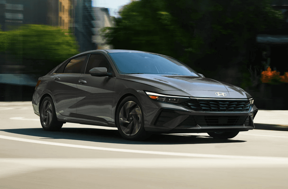 2025 Hyundai Elantra Hybrid in dark gray driving through an urban street with blurred city background.
