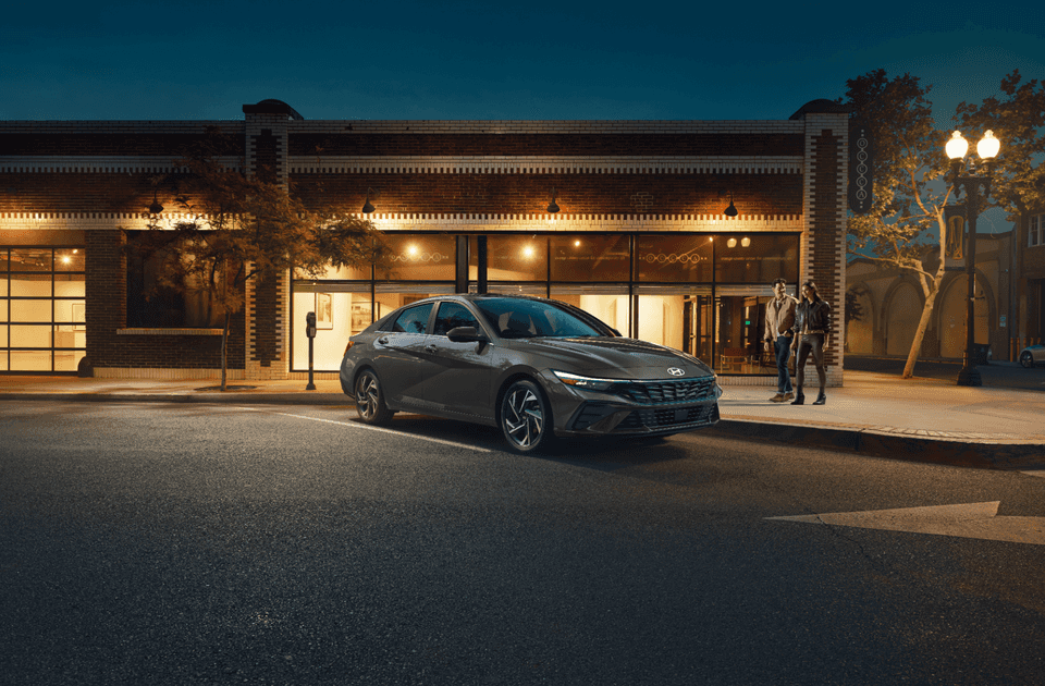 2025 Hyundai Elantra Hybrid parked on a city street at night in front of a well-lit brick building, with two people walking nearby.