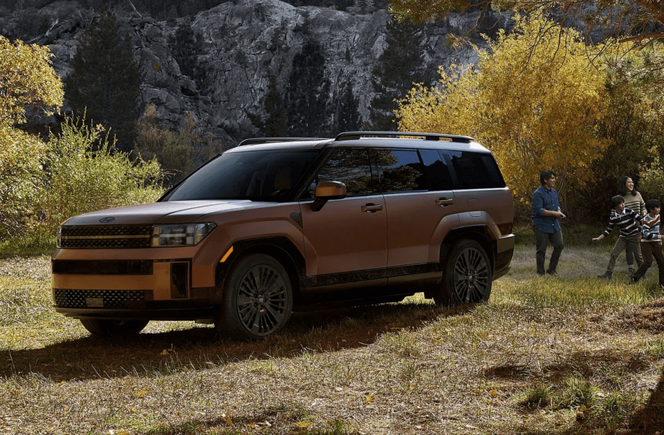2025 Hyundai Santa Fe Hybrid in bronze parked near a forest as a family plays nearby during fall.