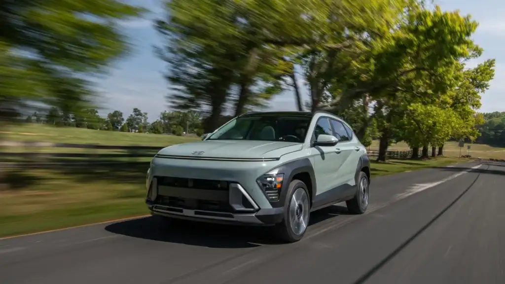 Light green 2026 Hyundai Kona compact SUV driving through scenic countryside highway.