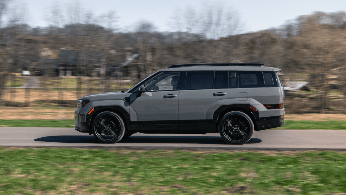 2026 Hyundai Santa Fe SUV driving in profile view on a paved road, showcasing bold body lines, black wheels, and rugged midsize styling.