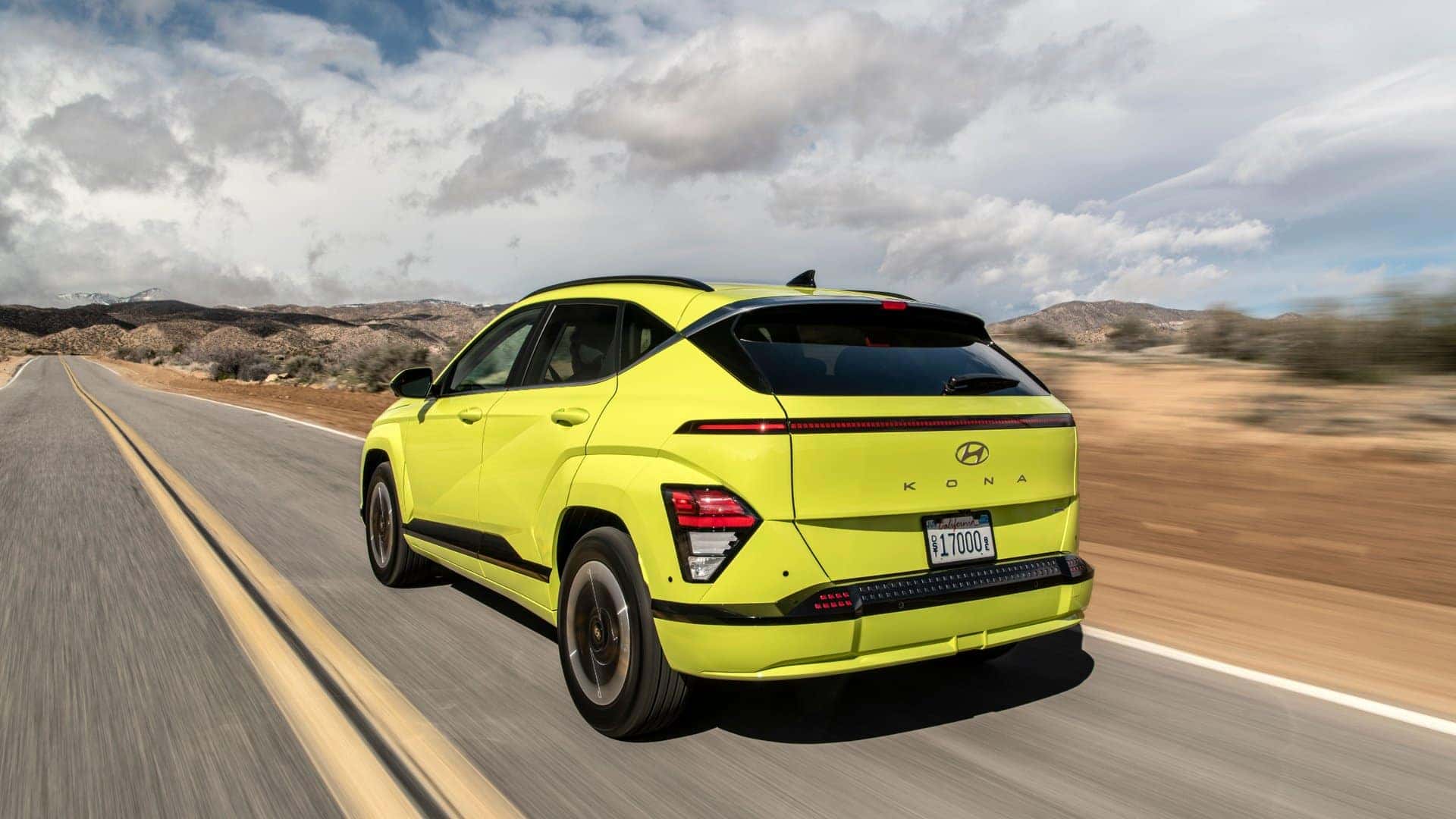 2025 Hyundai Kona Electric compact SUV driving on a desert highway with mountains in the background.