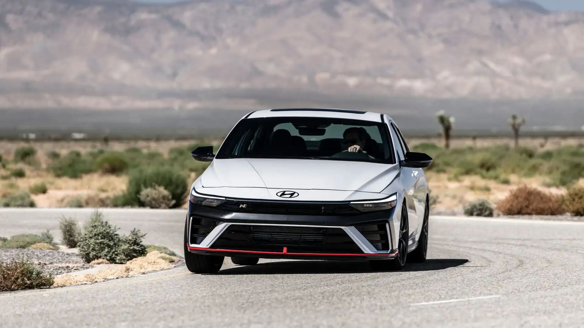 2026 Hyundai Elantra N white sport sedan driving on desert road with front grille and red accent trim.