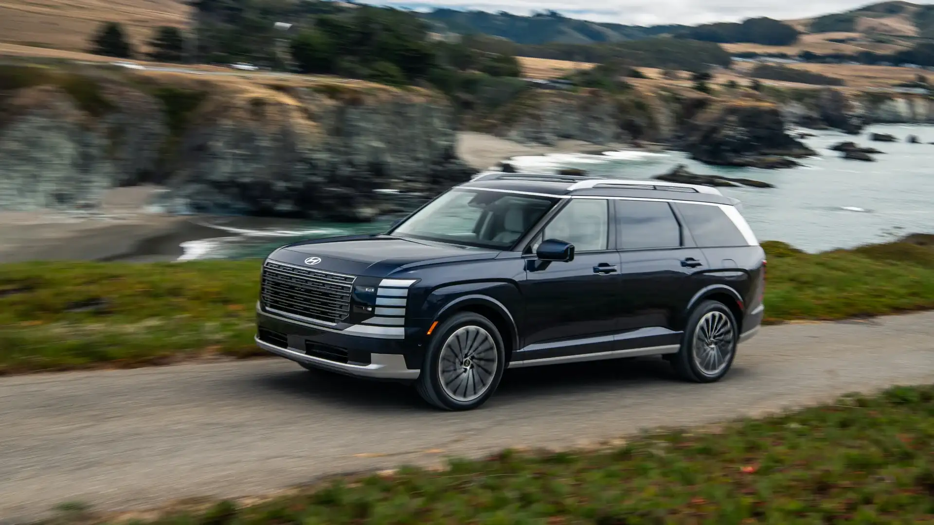2027 Hyundai Palisade black SUV driving along coastal road with ocean cliffs and mountain backdrop.