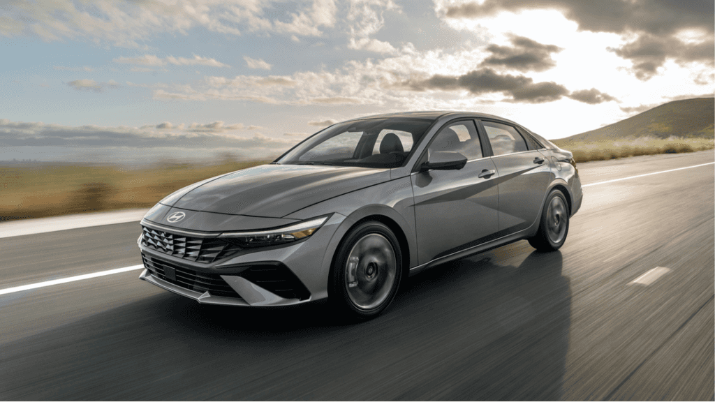 2025 Hyundai Elantra sedan in metallic gray driving on a scenic highway with hills and clouds in the background.