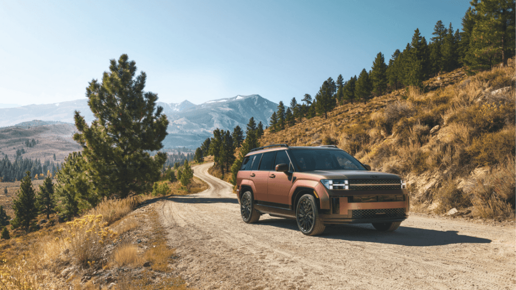 2025 Hyundai Santa Fe Hybrid SUV in a rugged bronze finish driving on a mountain dirt road surrounded by pine trees and scenic alpine views.
