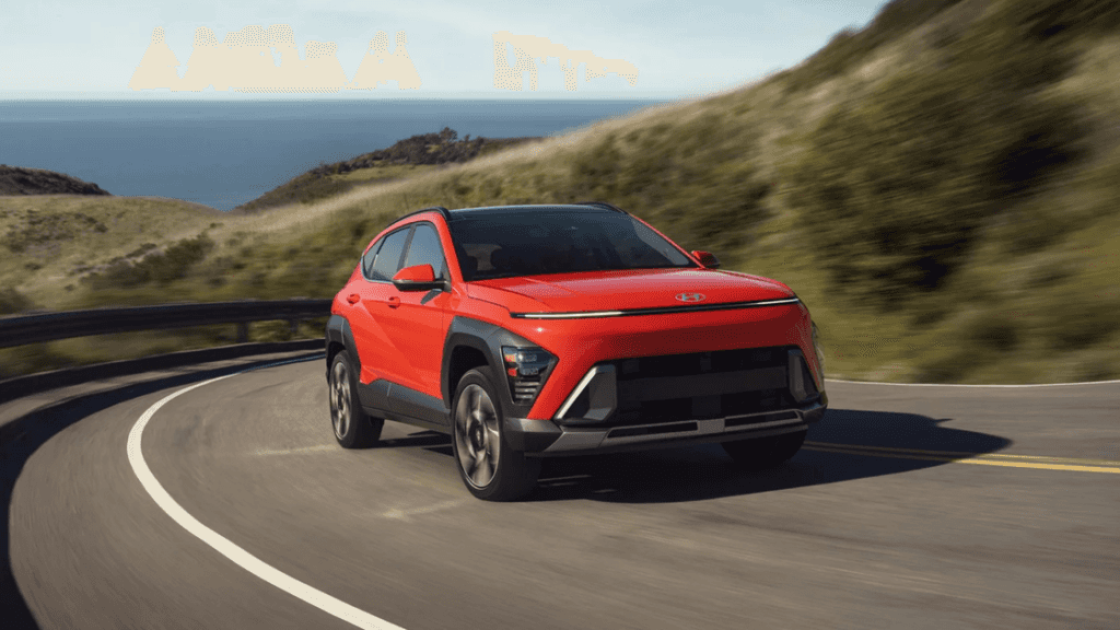 2025 Hyundai Kona in red driving along a coastal road with ocean and hills in the background.