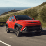 2025 Hyundai Kona in red driving along a coastal road with ocean and hills in the background.